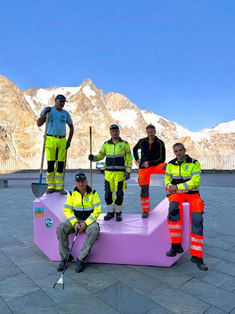 Five staff members in workwear pose on and beside a pink Enzi against an Alpine mountain backdrop.