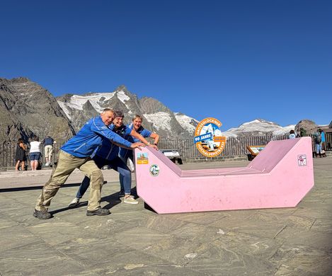 Three staff members pose on and beside a pink Enzi with the mountains of the Grossglockner High Alpine Road in the background.