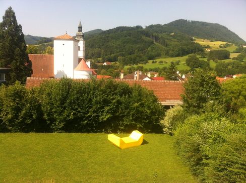 Ein gelbes Enzi-Sitzmöbel steht auf einer grünen Wiese mit Blick auf ein Dorf und bewaldete Hügel.