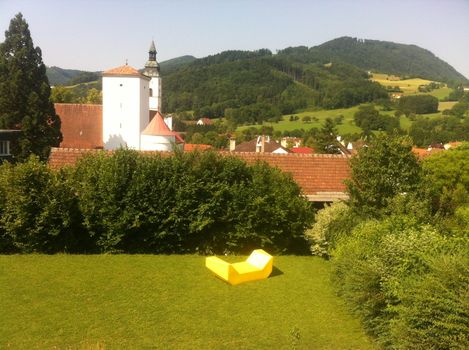 Ein gelbes Enzi-Sitzmöbel steht auf einer grünen Wiese mit Blick auf ein Dorf und bewaldete Hügel.
