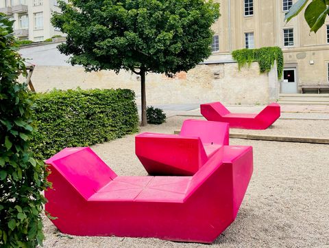 Mehrere pinke Enzi-Sitzmöbel stehen in einem begrünten Innenhof mit Bäumen und historischer Architektur.