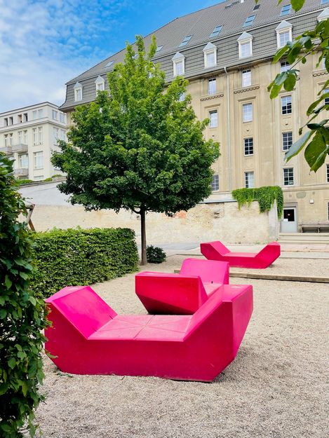 Mehrere pinke Enzi-Sitzmöbel stehen in einem begrünten Innenhof mit Bäumen und historischer Architektur.