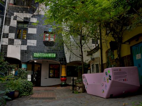 Rosa Enzi mit verschiedenen Stickern im begrünten Innenhof des Kunst Haus Wien, vor der charakteristischen schwarz-weißen Fassade.