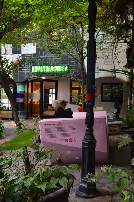 Zwei Personen sitzen auf einem rosa Enzi im begrünten Innenhof des Kunst Haus Wien, im Hintergrund der Eingang mit grün leuchtendem Schriftzug.