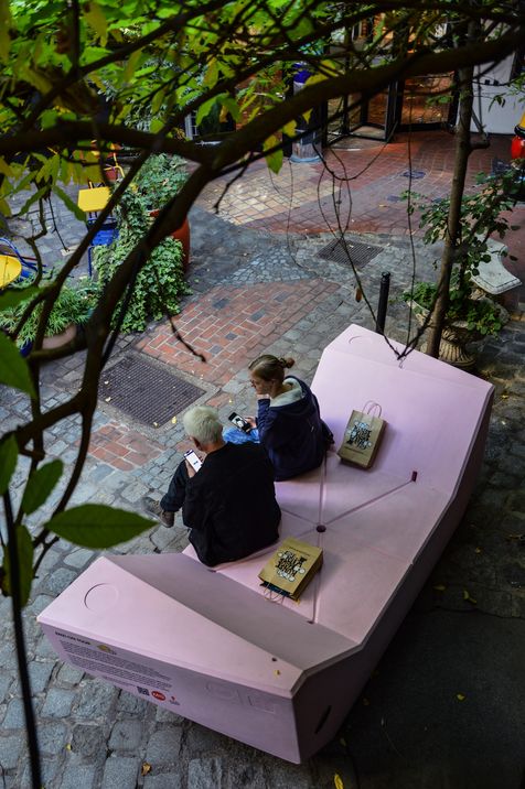 Zwei Personen sitzen auf einem rosa Enzi im Innenhof des Kunst Haus Wien und schauen auf ihre Smartphones.