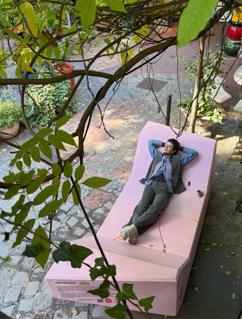 Eine Person liegt entspannt mit verschränkten Armen hinter dem Kopf auf einem rosa Enzi im begrünten Innenhof des Kunst Haus Wien.