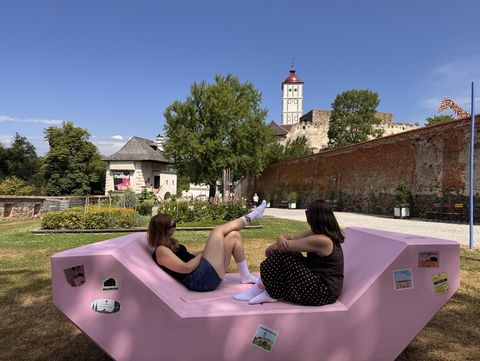 Zwei Frauen sitzen auf einem rosa Enzi im Schlosspark Schallaburg und genießen den Blick auf die Burgmauer und den Schlossturm. Im Hintergrund ragt eine Kunstinstallation in Form einer Giraffe über die Mauer.