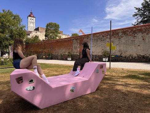 Ein rosa Enzi steht auf einer Wiese im Schlosspark Schallaburg vor einer historischen Burgmauer und dem Schlossturm. Das Möbel ist mit Aufklebern der „Enzi on Tour“-Stationen gestaltet.