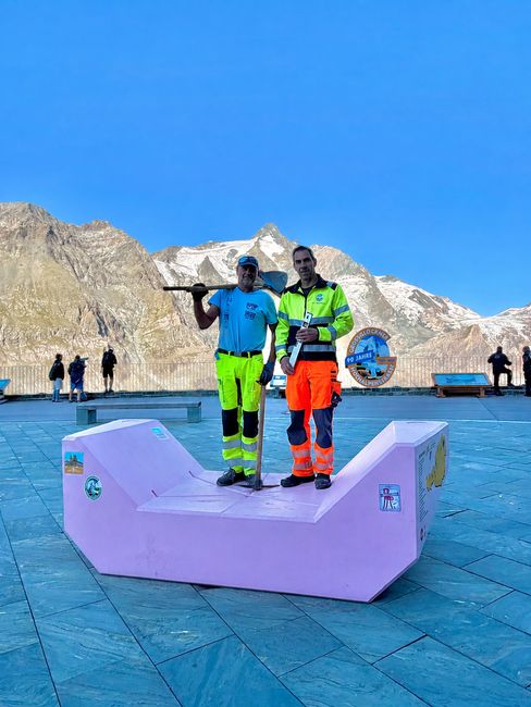 Two staff members with work equipment stand on a pink Enzi against the mountain scenery of the Grossglockner High Alpine Road.