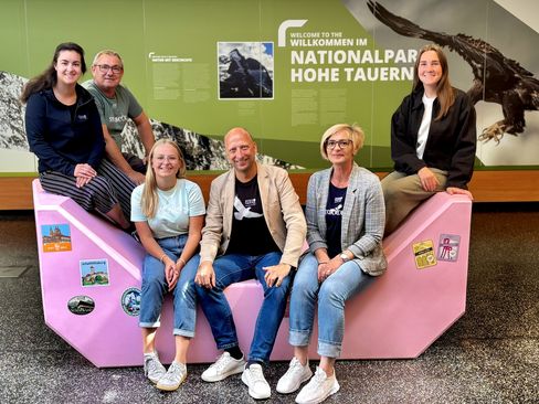 Sechs Mitarbeiter:innen des Nationalparks Hohe Tauern sitzen und stehen gemeinsam auf einem rosa Enzi im Besucherzentrum.