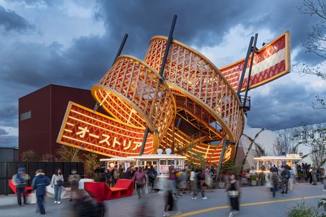 Rote Enzi-Sitzmöbel vor dem Österreich-Pavillon auf der Expo in Osaka, beleuchtete Holzstruktur mit geschwungenen Formen, viele Besucher:innen in Bewegung bei Dämmerung.
