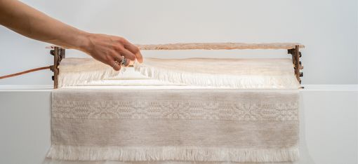 Hand mit Ringen hebt einen Holzrahmen über ein Webstück mit Fransen und feinem Muster