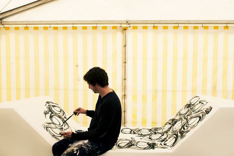 A person in dark clothing paints black circular patterns on a white bench. The background features yellow and white striped fabric, creating a creative, focused atmosphere.
