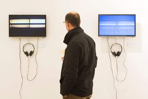 A man wearing glasses and a black jacket stands in front of two screens on a white wall, each screen has headphones hanging below it.