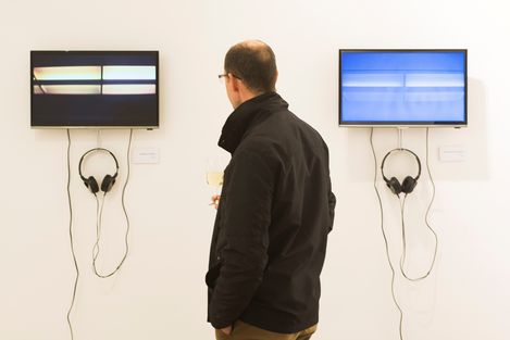 A man wearing glasses and a black jacket stands in front of two screens on a white wall, each screen has headphones hanging below it.