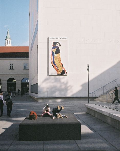 Exterior view of the Leopold Museum with an exhibition poster on the white façade; children playing on a black platform in the courtyard.