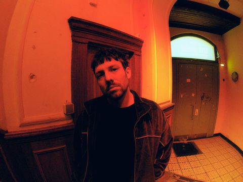 Person wearing a dark jacket standing in a hallway with tall walls, wood paneling, and a door with a transom window in the background.