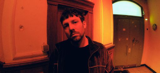 Person wearing a dark jacket standing in a hallway with tall walls, wood paneling, and a door with a transom window in the background.