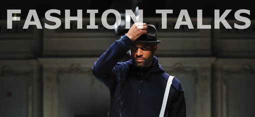 Man in dark clothing and hat walking on runway with audience on both sides, text "FASHION TALKS" and "WIENER FASHION WEEK" overlaid.