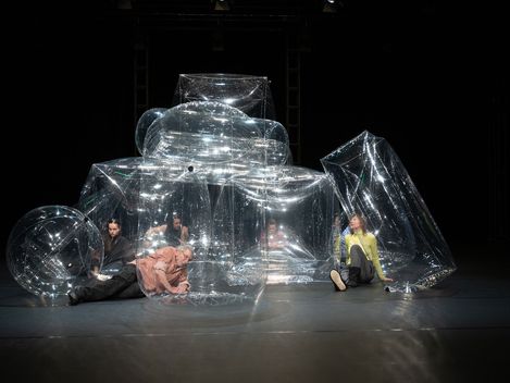 Die Performer*innen sitzen auf der Bühne inmitten transparenter, aufblasbarer Luftobjekte