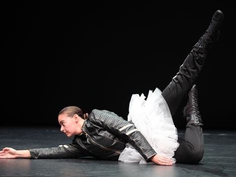 Doris Uhlich is performing a ballet pose while lying on the floor. She is wearing a tutu over her black leather outfit.
