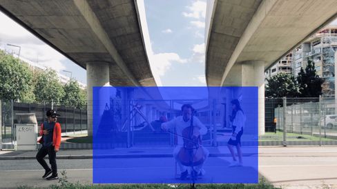 Under a highway bridge, a person is sitting playing a cello while another person in a red top and black cap stands to the left and a third person walks by on the right.