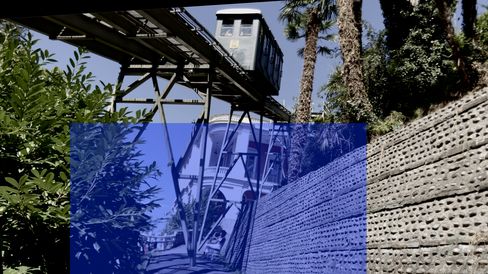 Suspended railway on steel supports above a path next to a perforated concrete wall and trees.