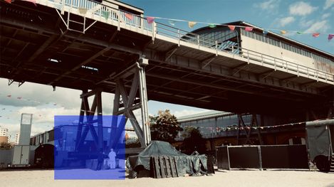 Under a high steel railway bridge stands a tarpaulin-covered structure next to a fence and several trucks with buildings and colorful pennant strings in the background.