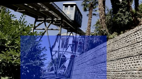 Suspended railway on steel supports above a path next to a perforated concrete wall and trees.
