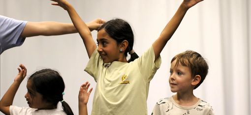 Zu sehen sind 3 Kinder, 2 Mädchen und ein Junge, in der Mitte ein großes Mädchen, das die Arme in die Höhe streckt und nach rechts schaut, daneben ein kleineres Mädchen, dass die Arme angewinkelt nach oben hält, auf der anderen Seite ein Junge, der in dieselbe Richtung blickt. Im Hintergrund ist der Arm einer größeren Person zu sehen, vielleicht von der Workshopleitung.
