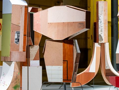 Wooden geometric sculptures with varied textures and colors stand on black metal legs in a brightly lit gallery, creating a modern and abstract feel.