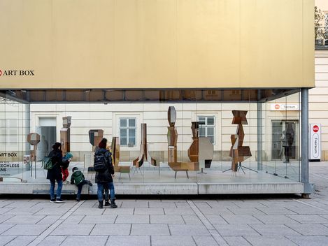 A family admires the 'Speechless Signs' art installation by Fabian Seiz in an MQ Art Box in Vienna, Austria. The glass box showcases abstract sculptures and draws in curious onlookers.