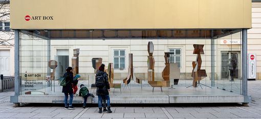 A family admires the 'Speechless Signs' art installation by Fabian Seiz in an MQ Art Box in Vienna, Austria. The glass box showcases abstract sculptures and draws in curious onlookers.