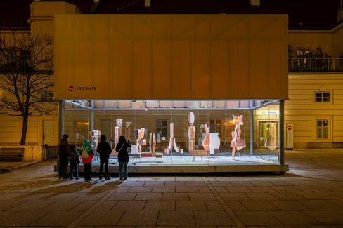 Group of people viewing an illuminated display box at night containing abstract vertical sculptures made of wood and paper.