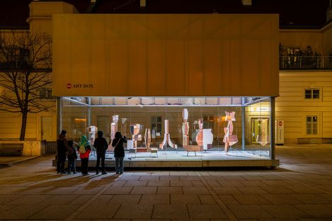 Group of people viewing an illuminated display box at night containing abstract vertical sculptures made of wood and paper.