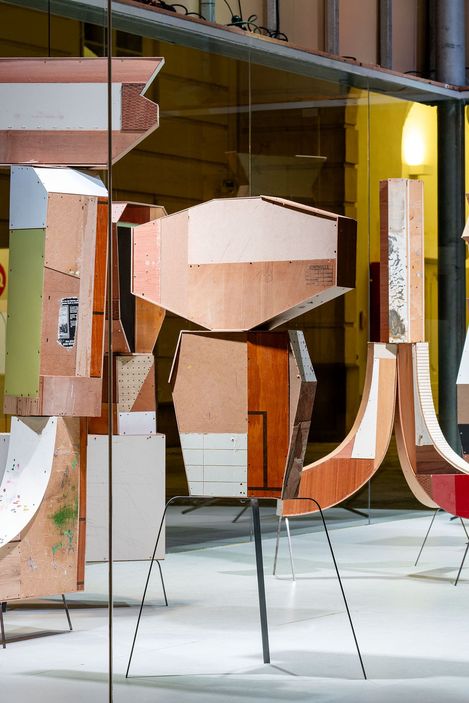 Wooden geometric sculptures with varied textures and colors stand on black metal legs in a brightly lit gallery, creating a modern and abstract feel.