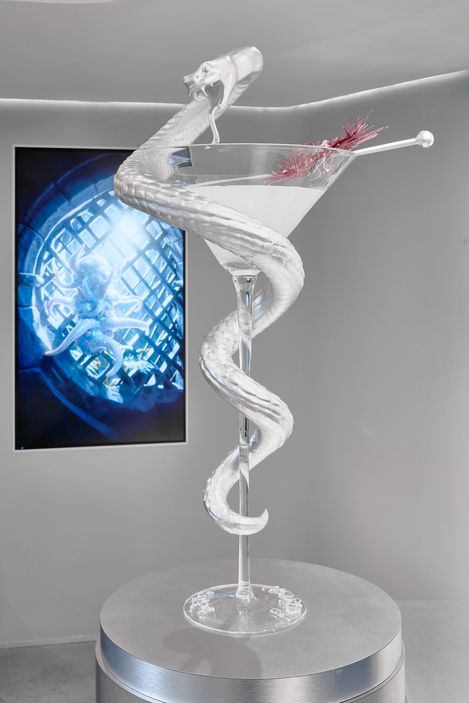 Sculpture of a large martini glass with a snake coiled around it and a cocktail pick with red fruit pieces in an exhibition space
