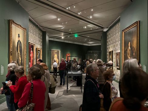 Visitors in a museum gallery with several framed portrait paintings on the walls and a display case in the center.