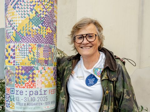 Frau mit Brille, weißem T-Shirt und Camouflage-Jacke lehnt an einem mit buntem Muster und Text beklebten runden Pfeiler und lächelt in die Kamera
