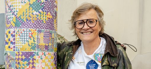 Frau mit Brille, weißem T-Shirt und Camouflage-Jacke lehnt an einem mit buntem Muster und Text beklebten runden Pfeiler und lächelt in die Kamera