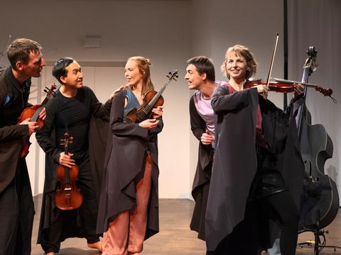 Five people in dark costumes on a stage, four holding violins, one standing next to a double bass.