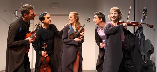 Five people in dark costumes on a stage, four holding violins, one standing next to a double bass.
