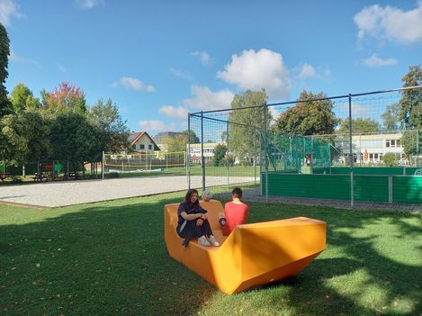 Zwei Personen sitzen auf einem gelben Enzo auf einer Wiese neben einem Sportplatz und nutzen das Sitzmöbel als Treffpunkt.