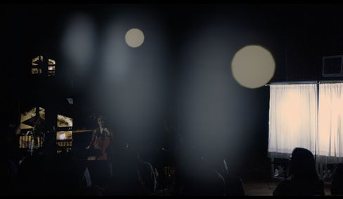 Dark room with a person playing a cello and several light sources including two large round light spots and a lit window with curtains.