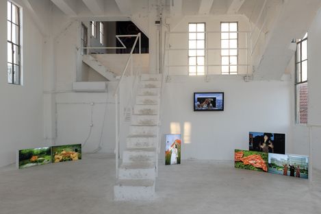 Interior of a white gallery space with a central staircase and multiple photo screens placed on the floor.