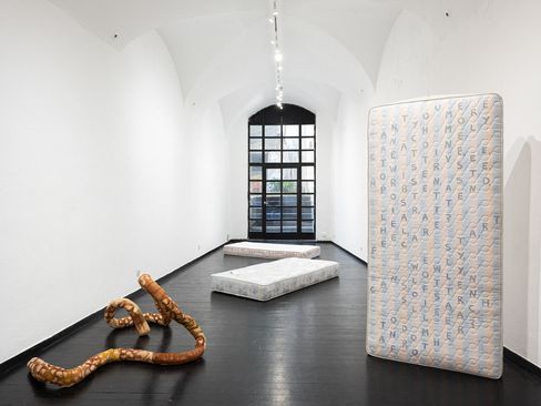 Three mattresses in a white vaulted room with a black floor, one mattress standing upright displaying a pattern of letters, in front a sculptural curved wooden form.