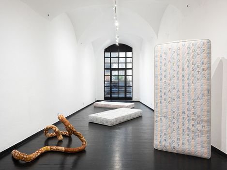 Three mattresses in a white vaulted room with a black floor, one mattress standing upright displaying a pattern of letters, in front a sculptural curved wooden form.