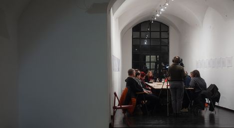 Several people sit around a long table in a white, high-ceilinged room with arched ceiling and large windows, one person stands with a camera.
