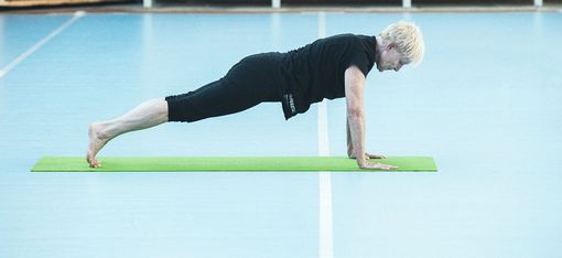 Person with short light hair wearing black sportswear holding a plank position on a green yoga mat in a gym with a blue floor