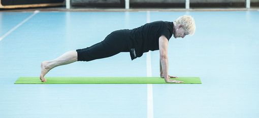 Person mit kurzen hellen Haaren in schwarzer Sportkleidung hält eine Plank-Position auf einer grünen Yogamatte in einer Halle mit blauem Boden